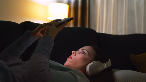 Mujer-Con-Auriculares-Inalámbricos-Recostada-En-Un-Sofá-En-Casa-Por-La-Noche-Transmitiendo-O-Mirando-Contenido-En-Línea-En-Un-Teléfono-Móvil-1